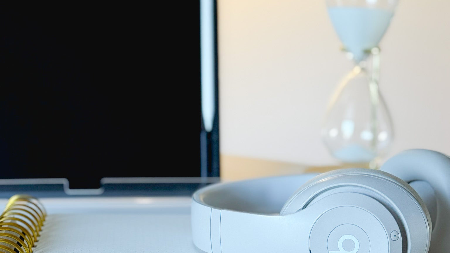 White Beats headphones on a desk with a laptop, notebook, and sand timer in the background.